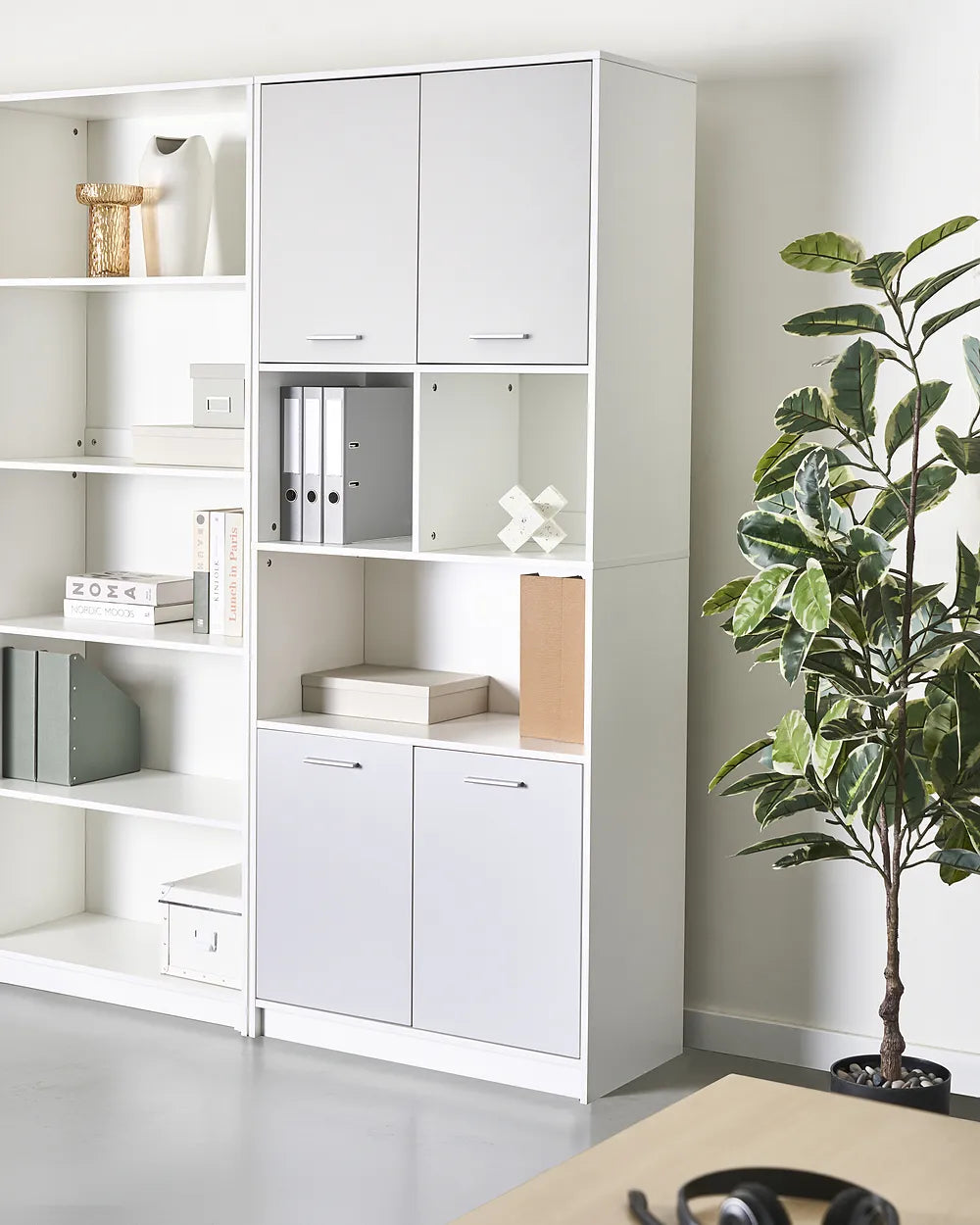 A tall white modular storage cabinet with open shelves and two lower doors, shown in a bright office-like room next to a potted plant. The cabinet is styled with binders, boxes, and decor.