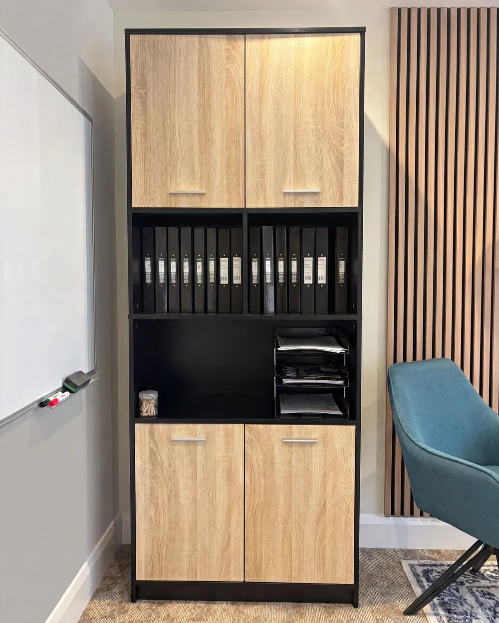 A tall black-framed office storage cabinet with light oak wood grain doors and open shelves, shown in a home office setting next to a whiteboard, wood slat wall, and teal chair.