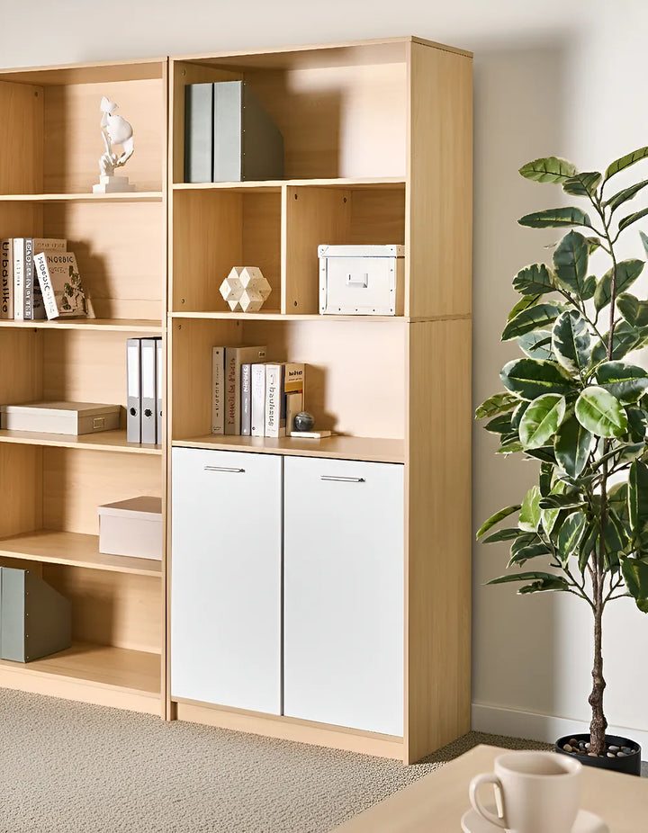 A tall light wood and white storage bookcase with open shelves and two cabinet doors, styled in a home office next to a potted plant.