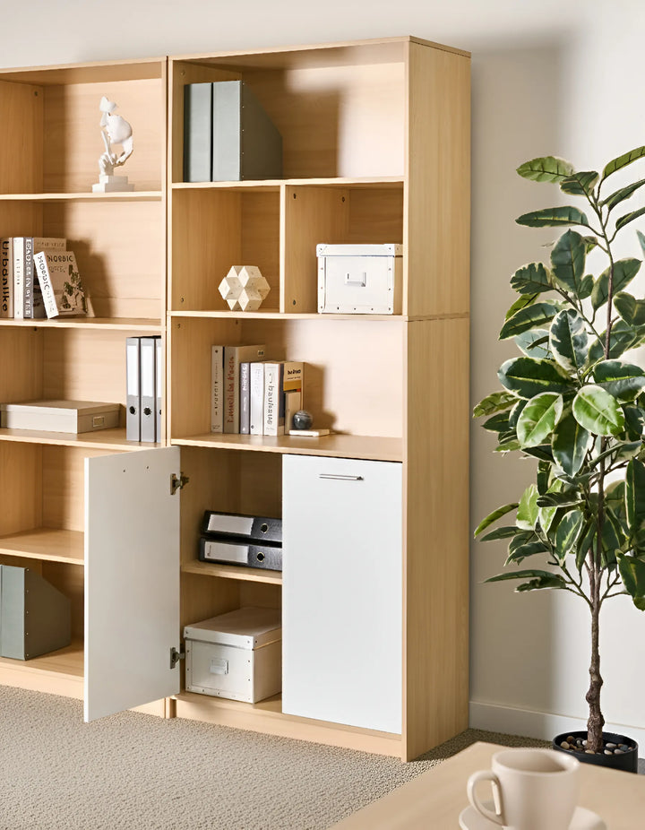 A light wood and white modular bookcase with open shelves and two cabinet doors, styled with books, binders, and boxes in a bright room next to a potted plant.