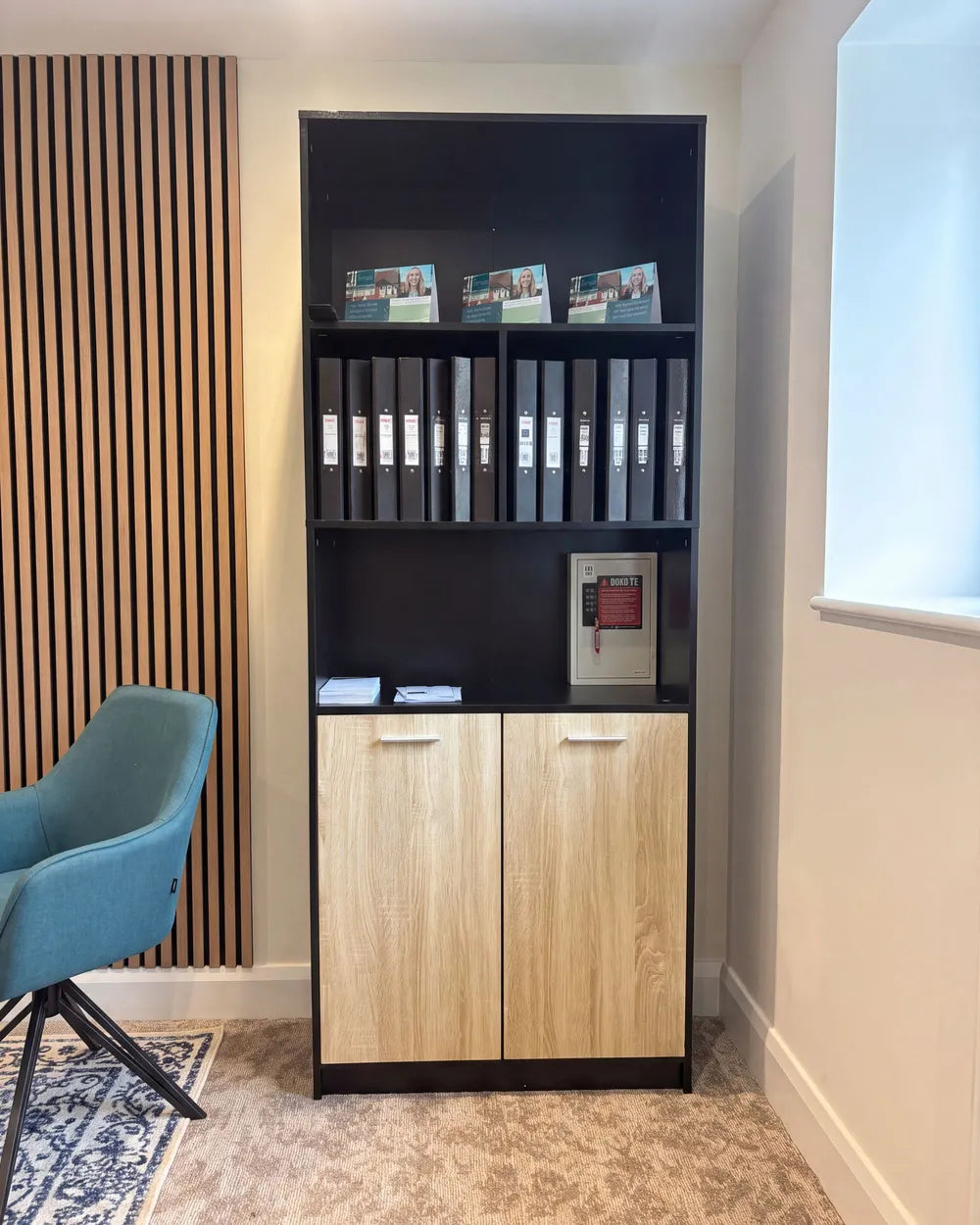 A tall black office shelving unit with two light wood cabinet doors at the bottom, shown in a home office setting next to a blue chair and slatted wood wall paneling.