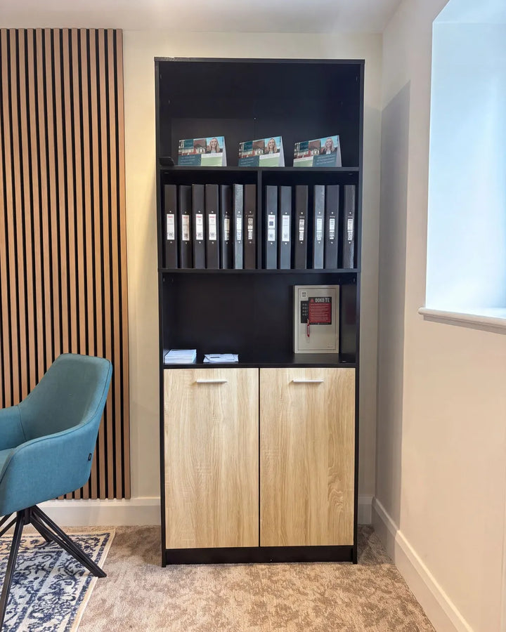 A tall black office shelving unit with two light wood cabinet doors at the bottom, shown in a home office setting next to a blue chair and slatted wood wall paneling.