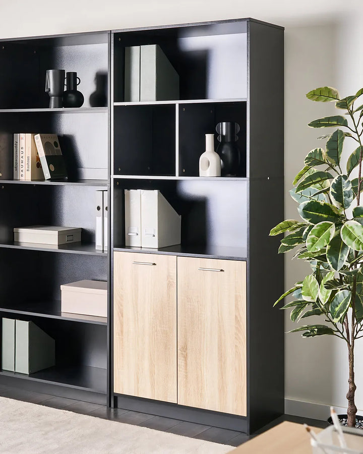 A tall black modular bookcase with two light wood cabinet doors at the bottom, styled with books, vases, and boxes in a modern room next to a potted plant.