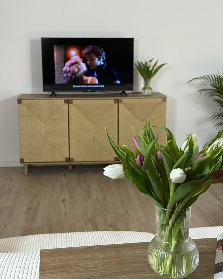 A modern wooden TV stand cabinet with a geometric patterned door design, holding a television in a living room setting with a vase of tulips in the foreground.