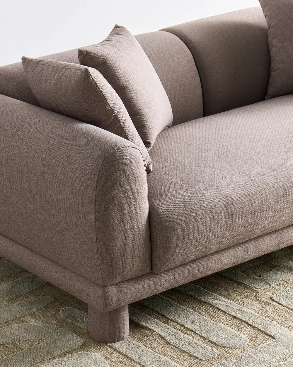 A close-up of a modern upholstered sofa in a warm taupe-beige fabric, featuring curved armrests, matching back cushions, and a textured patterned rug underneath.