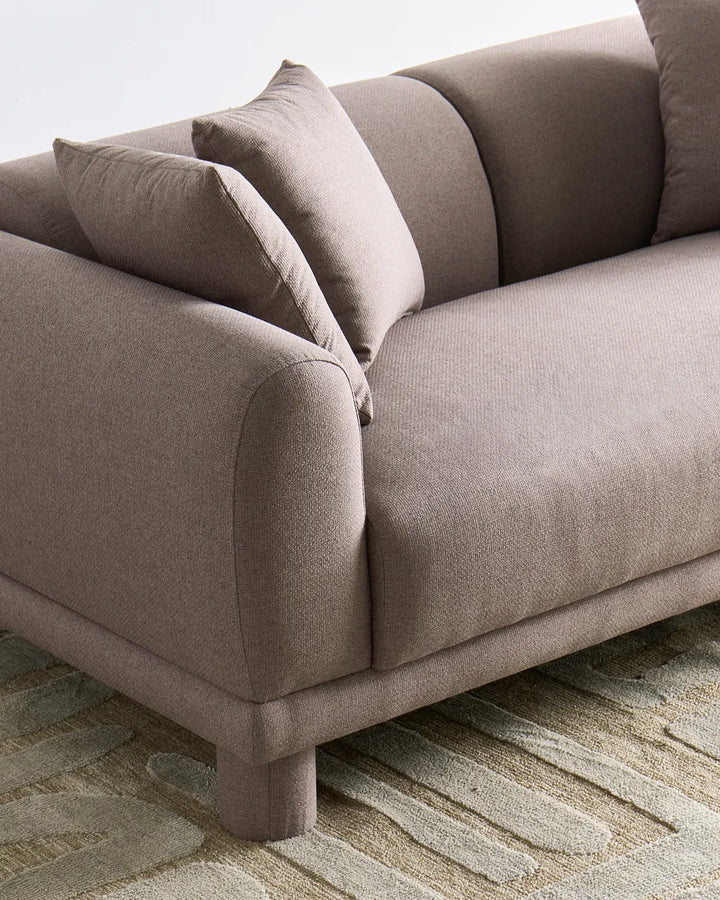 A close-up of a modern upholstered sofa in a warm taupe-beige fabric, featuring curved armrests, matching back cushions, and a textured patterned rug underneath.