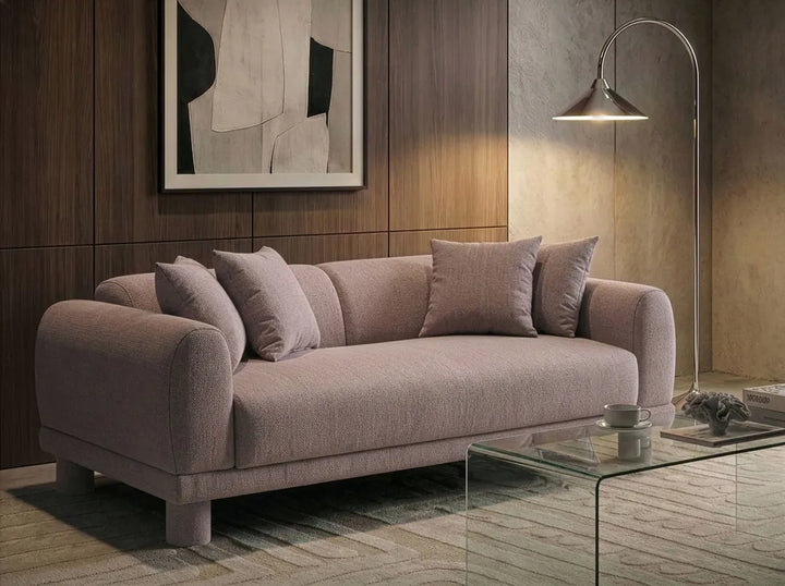 A modern three-seater sofa upholstered in a textured dusty mauve-pink fabric, styled in a contemporary living room with a glass coffee table, floor lamp, and wood-paneled wall.