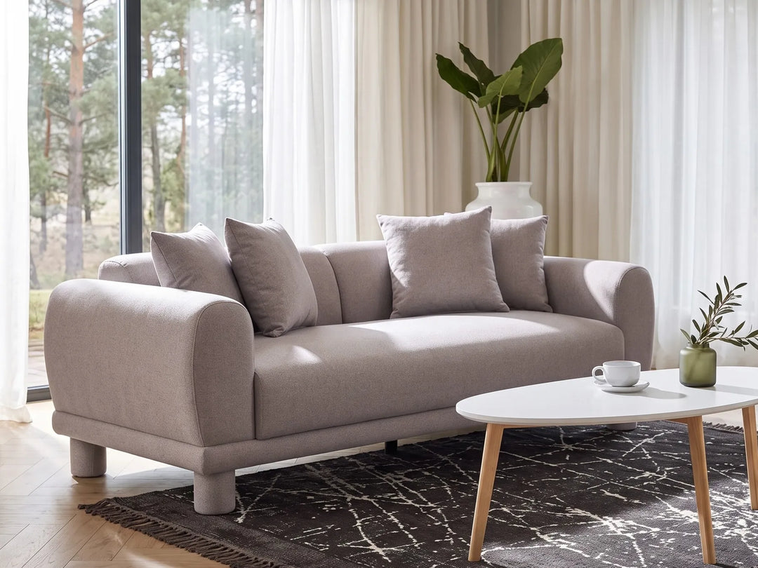 A modern beige fabric sofa with rounded arms and four matching throw pillows, styled in a bright living room with a white oval coffee table, dark patterned rug, and a large potted plant in the background.