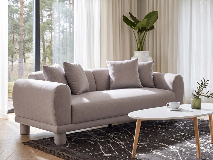 A modern beige fabric sofa with rounded arms and four matching throw pillows, styled in a bright living room with a white oval coffee table, dark patterned rug, and a large potted plant in the background.