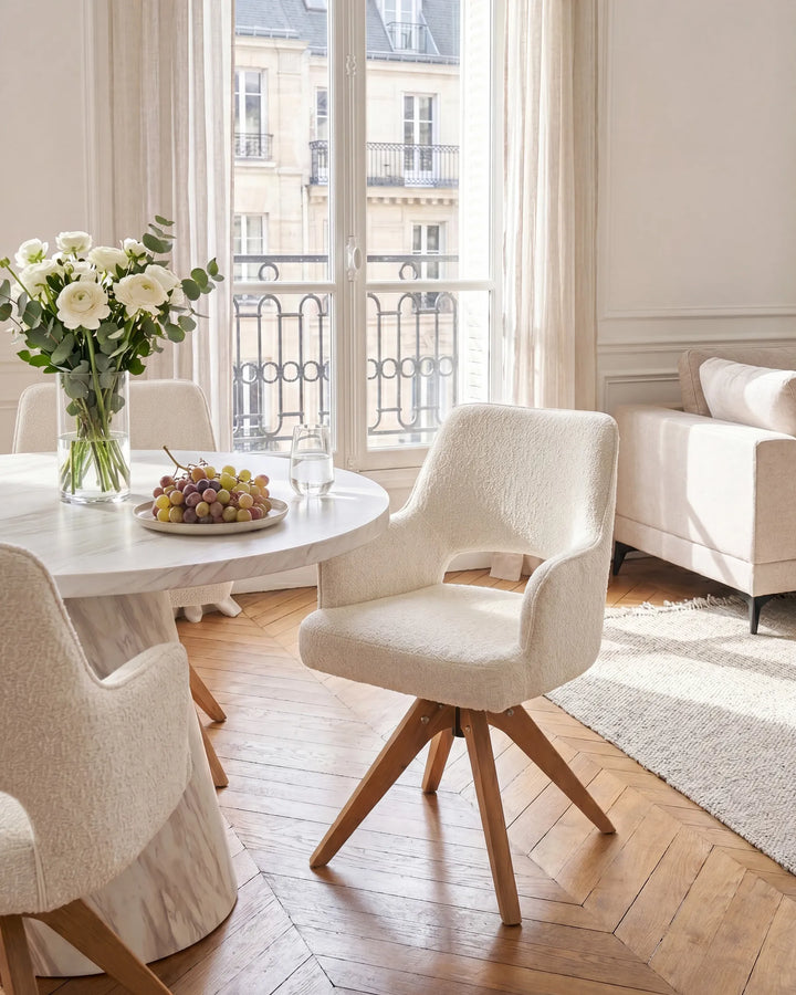 A modern dining area featuring textured cream bouclé swivel chairs with light wood legs, set around a round white table on a herringbone wood floor. Large windows in the background offer a view of Parisian buildings.