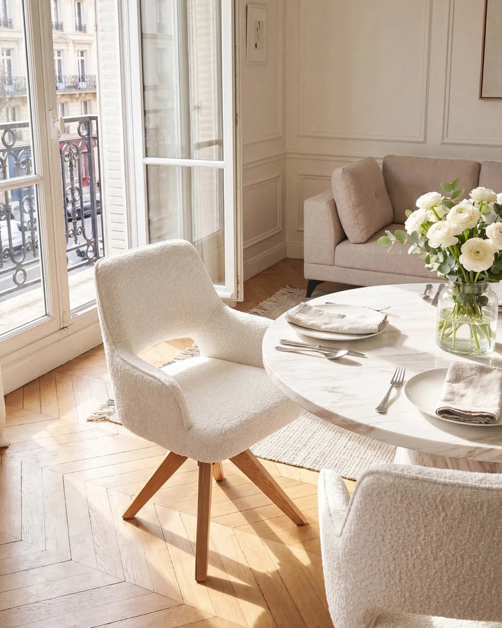 A modern dining room scene featuring a round white marble table set with white plates, silverware, and a vase of white roses, surrounded by textured cream boucle chairs. In the background, a light beige sofa sits in a sunlit room with white paneled walls and a balcony view.