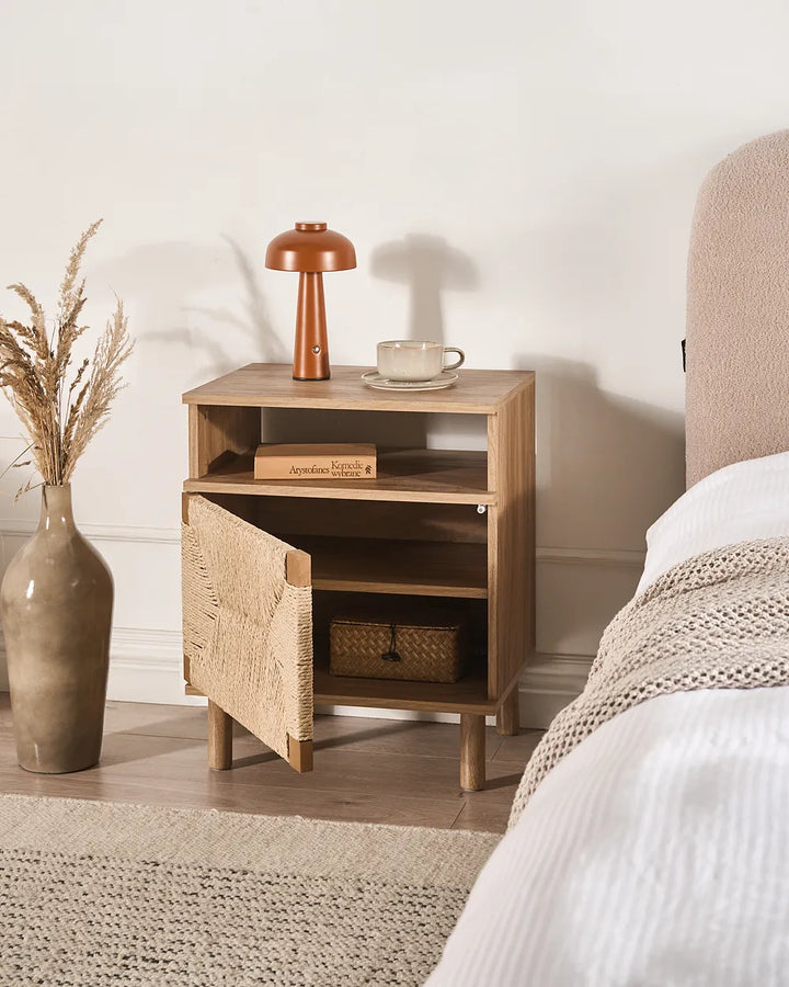 A light natural wood nightstand with an open door, styled in a bedroom next to a bed with white bedding and a beige headboard. The nightstand holds a copper-colored table lamp, a white coffee cup, and a book, with a textured vase of dried grass on the floor nearby.