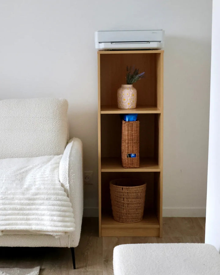 A tall, narrow light wood shelving unit with three open compartments, styled with a small vase, a woven basket, and a storage bin, set against a neutral wall next to a white textured armchair.