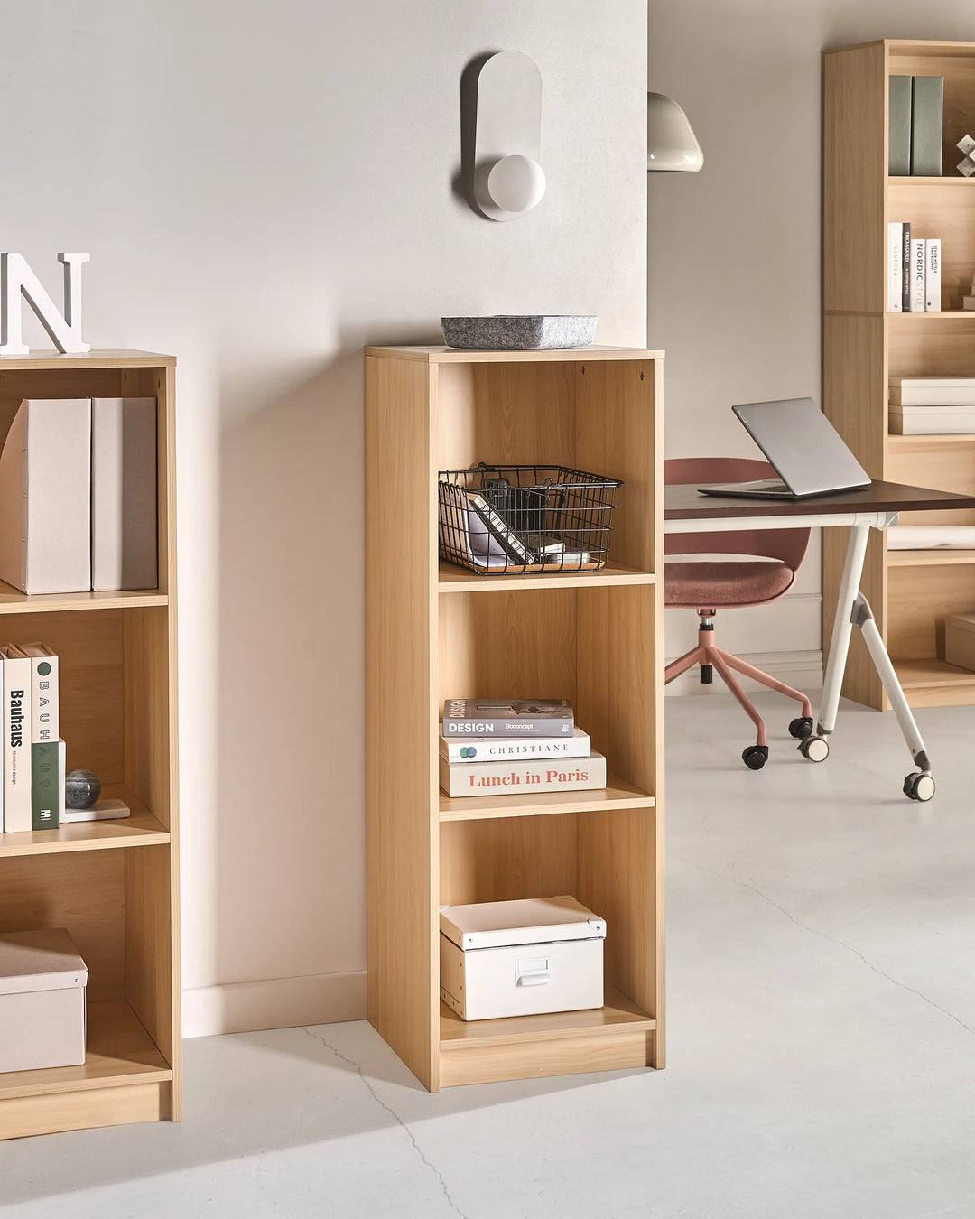 A tall, narrow light wood bookcase with four open shelves stands in a modern home office setting next to a larger matching bookcase. The shelves are styled with books, storage boxes, a wire basket, and a small bowl.