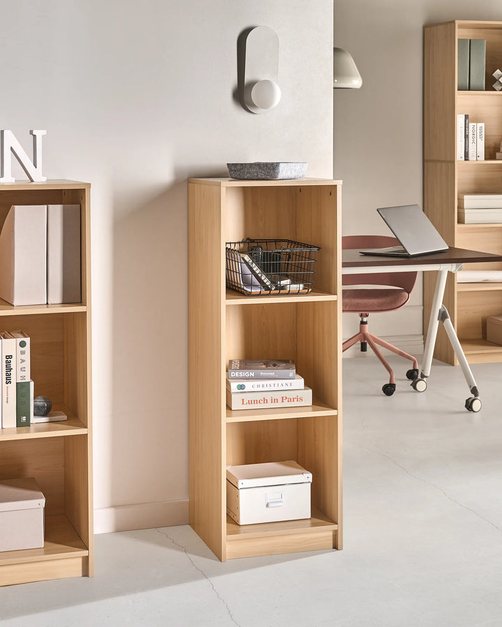 A tall, narrow light wood bookcase with four open shelves stands in a modern home office setting next to a larger matching bookcase. The shelves are styled with books, storage boxes, a wire basket, and a small bowl.
