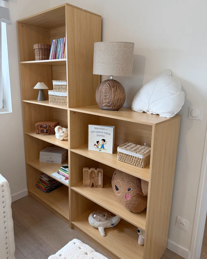 A light natural wood modular bookcase with six open shelves, styled with books, baskets, a lamp, a white cloud pillow, and plush toys in a bright room.
