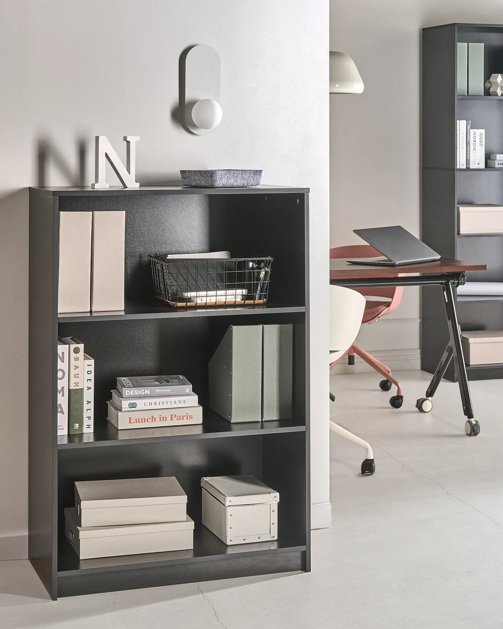 A tall, dark gray three-tier bookcase styled with books, storage boxes, and a wire basket in a modern office setting.