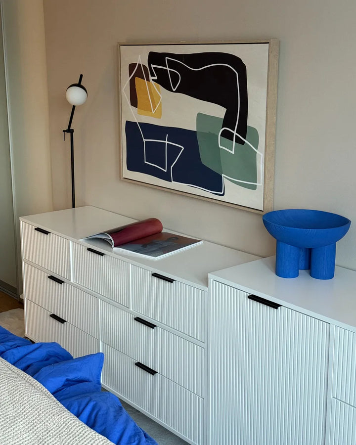 A modern bedroom scene featuring a large abstract geometric wall art print with black, navy, green, and yellow shapes, hung above a white ribbed dresser with black handles.