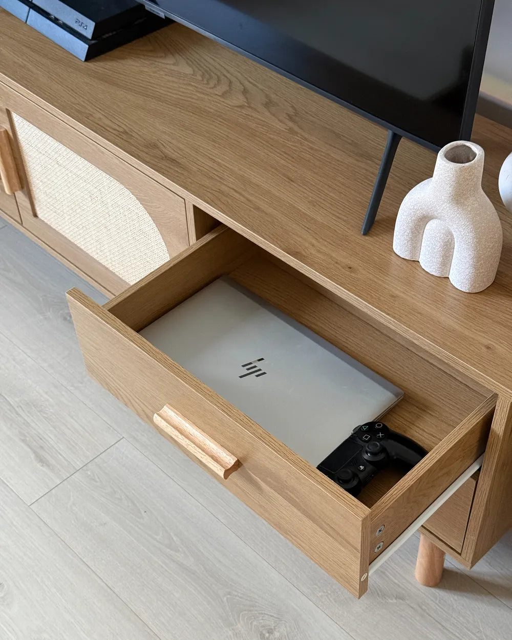 A close-up of a light oak TV stand or console cabinet with a drawer pulled open, showing a silver laptop and a black game controller inside. The cabinet has woven cane panels on the side, a light wood handle, and is styled in a modern room setting with a black TV and white ceramic vases on top.