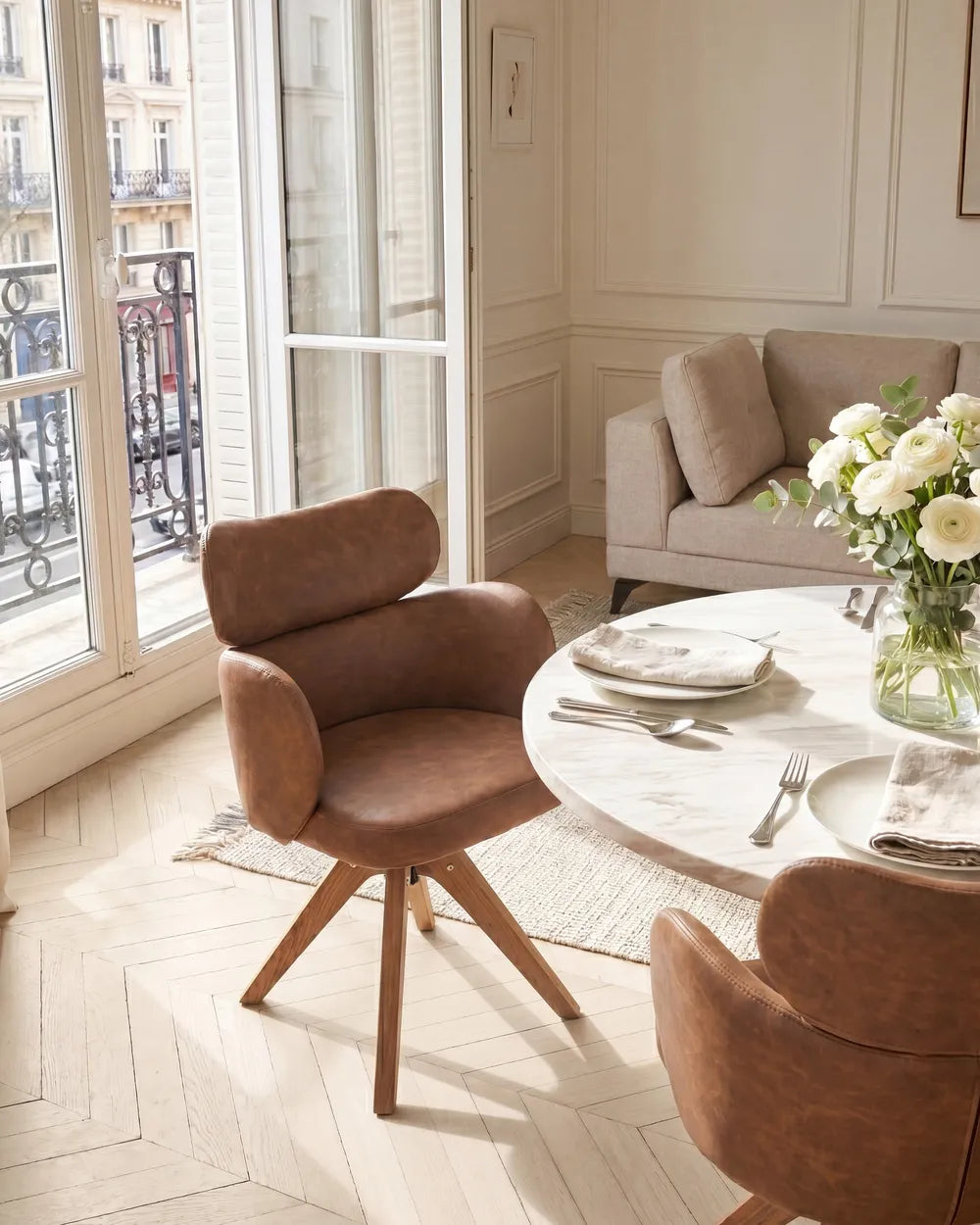 A warm brown upholstered dining chair with a swivel base and wooden legs, shown in a bright, modern dining room setting next to a marble table with white flowers.