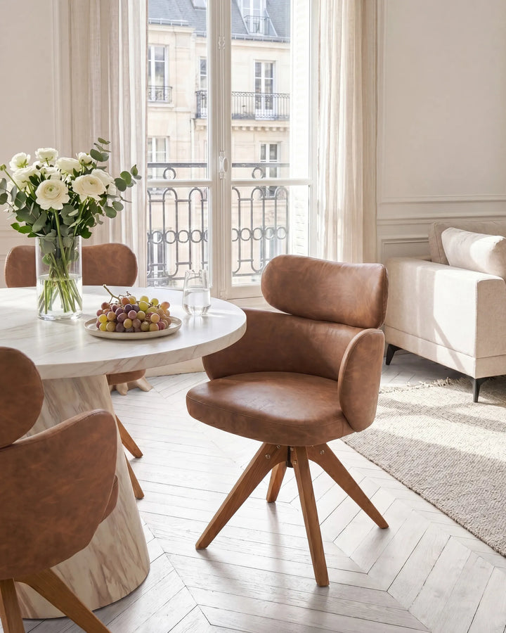 A modern brown leather swivel dining chair with wooden legs in a bright, elegant dining room setting with a round marble table and a window overlooking Parisian buildings.