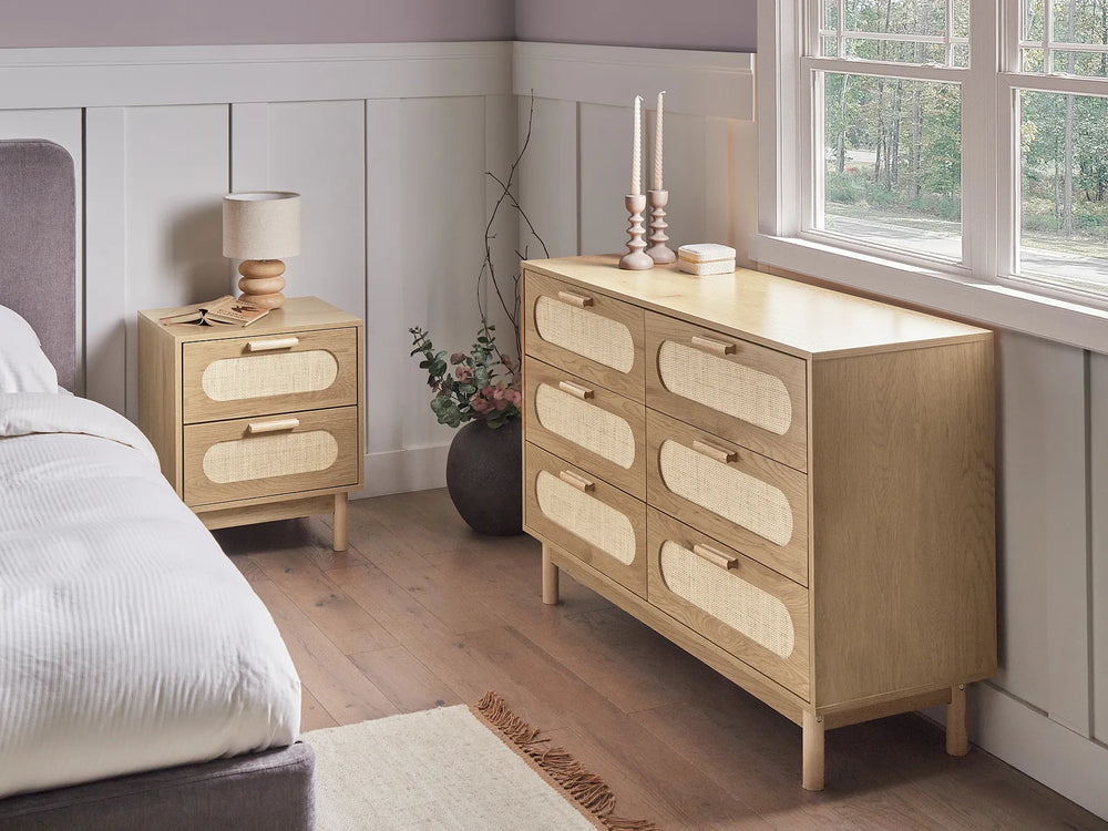 A light natural wood six-drawer dresser with woven cane drawer fronts stands beside a matching two-drawer nightstand in a styled bedroom setting with a bed, window, and wood flooring.