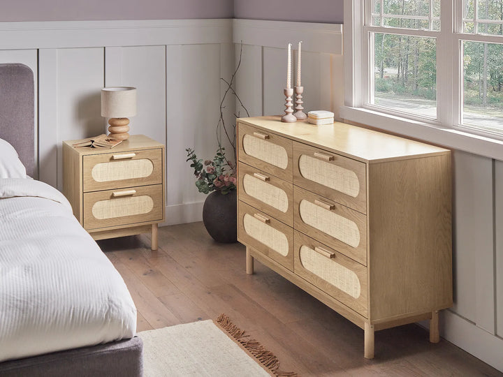 A light natural wood six-drawer dresser with woven cane drawer fronts stands beside a matching two-drawer nightstand in a styled bedroom setting with a bed, window, and wood flooring.