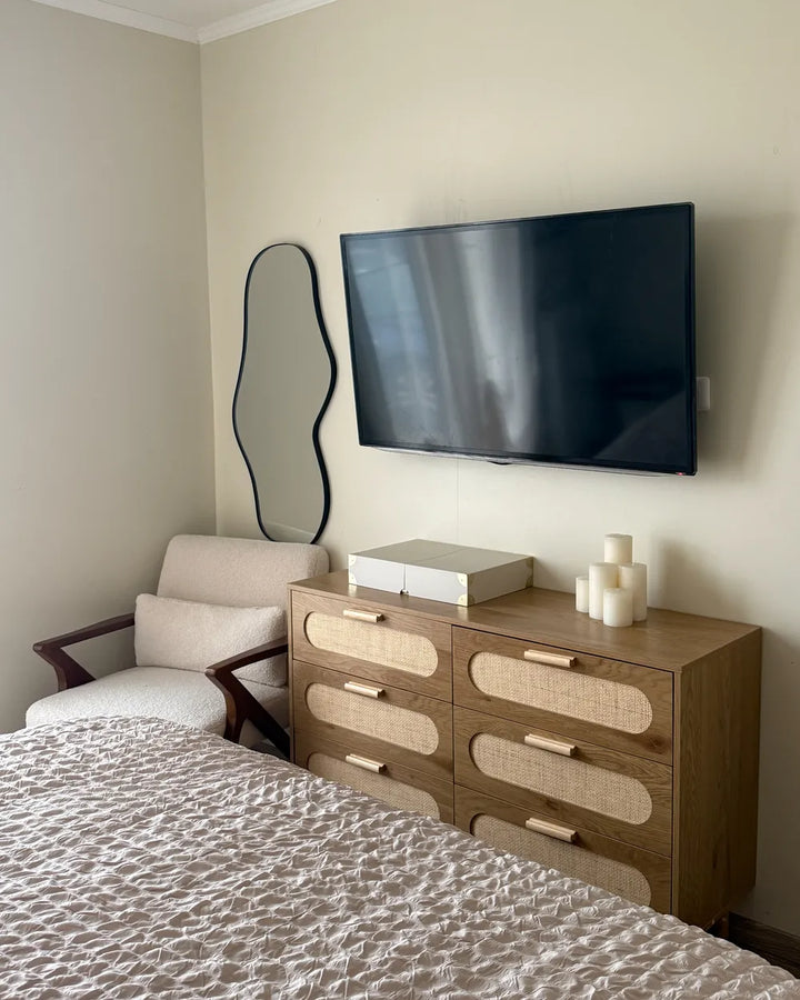 A mid-century modern bedroom scene featuring a wooden dresser with woven cane drawer fronts, a black wall-mounted TV, a wavy black-framed mirror, and a beige armchair.