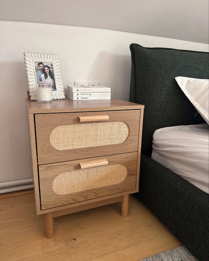 A two-drawer wooden nightstand with woven cane panels on the front and light wood drawer pulls, styled next to a bed with a dark green headboard. The nightstand has a white picture frame and a candle on top.