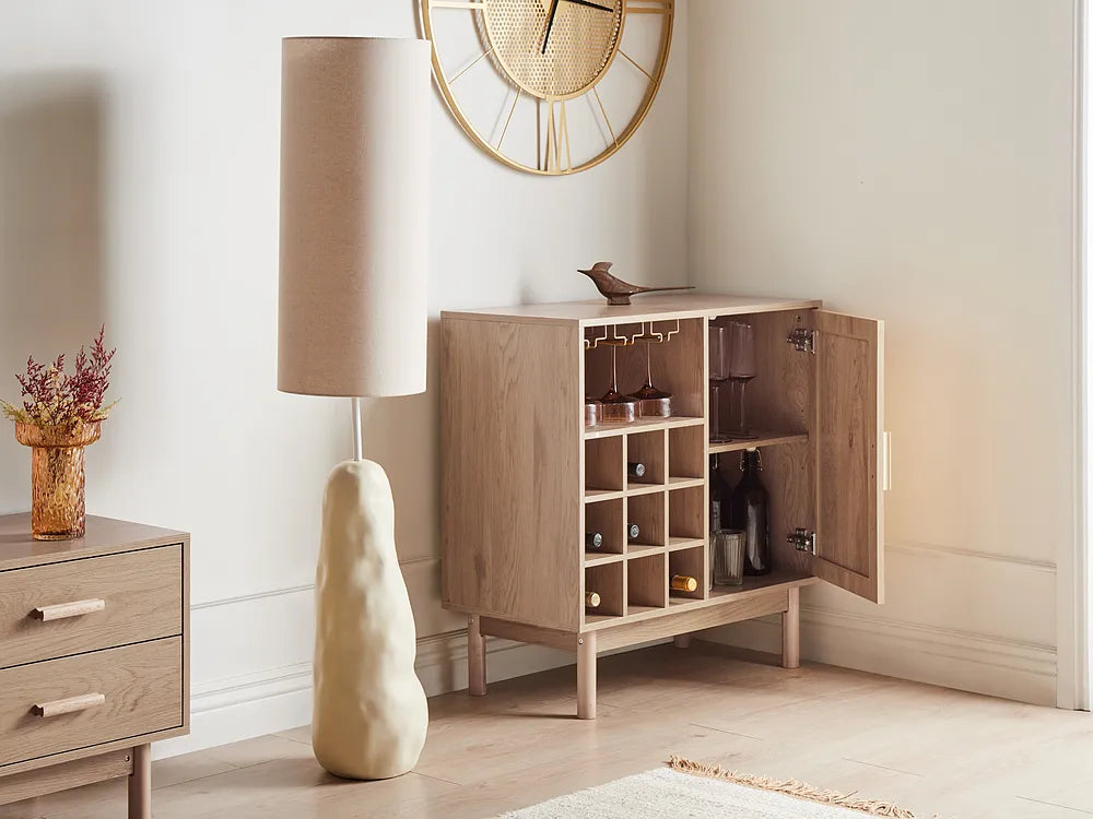 A light oak wood bar cabinet with an open door, showcasing wine bottles and glassware inside, styled in a modern room next to a floor lamp and a small side table.
