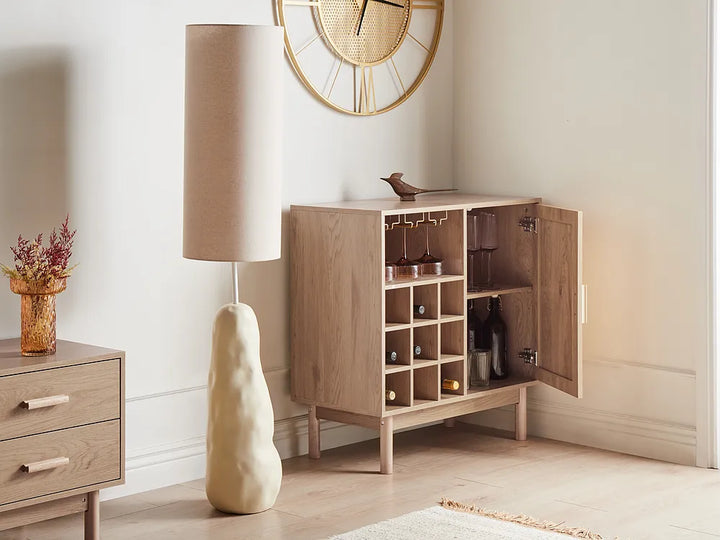 A light oak wood bar cabinet with an open door, showcasing wine bottles and glassware inside, styled in a modern room next to a floor lamp and a small side table.