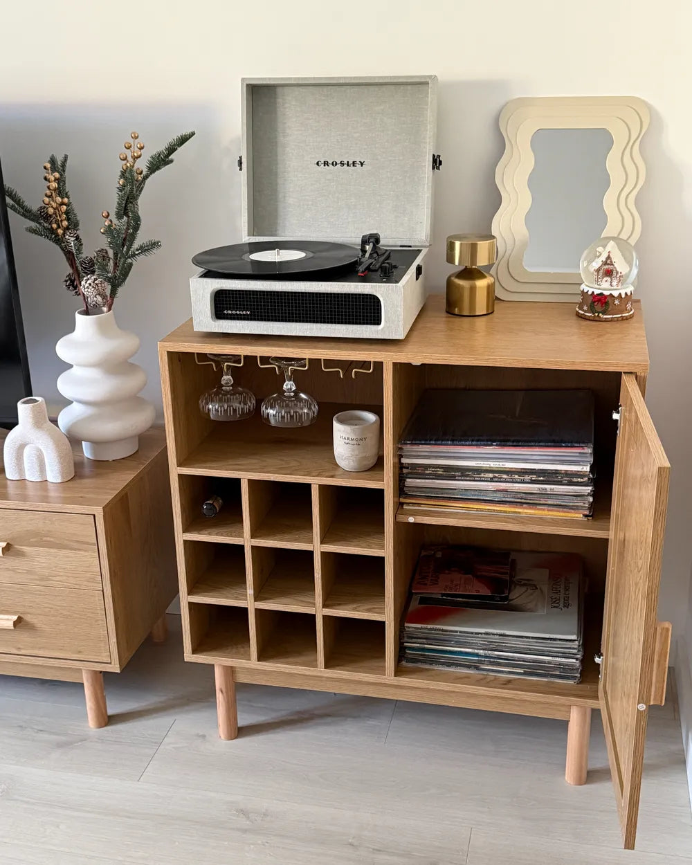 A mid-century modern wooden record cabinet with an open door, styled with a Crosley record player, vinyl records, glassware, and decorative accessories in a bright room.