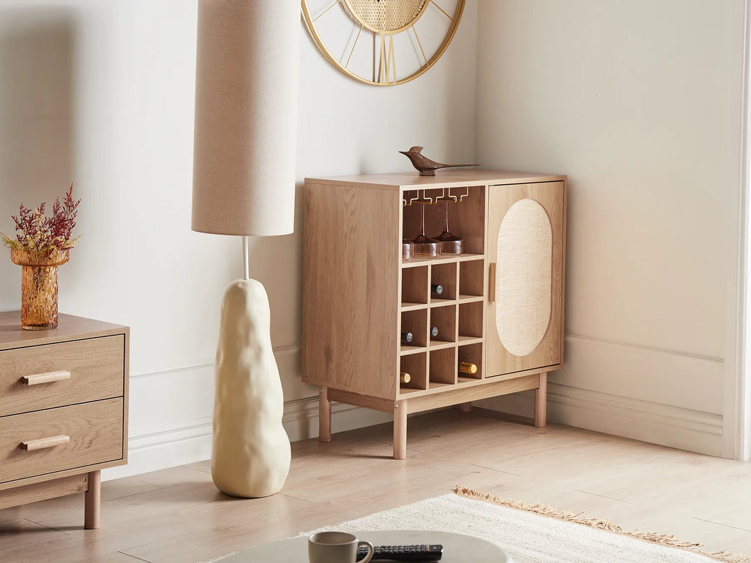 A light natural wood bar cart with a wine bottle rack, hanging glass hooks, and a round woven rattan door panel, styled in a modern minimalist room next to a matching wood nightstand and a textured cream floor lamp.