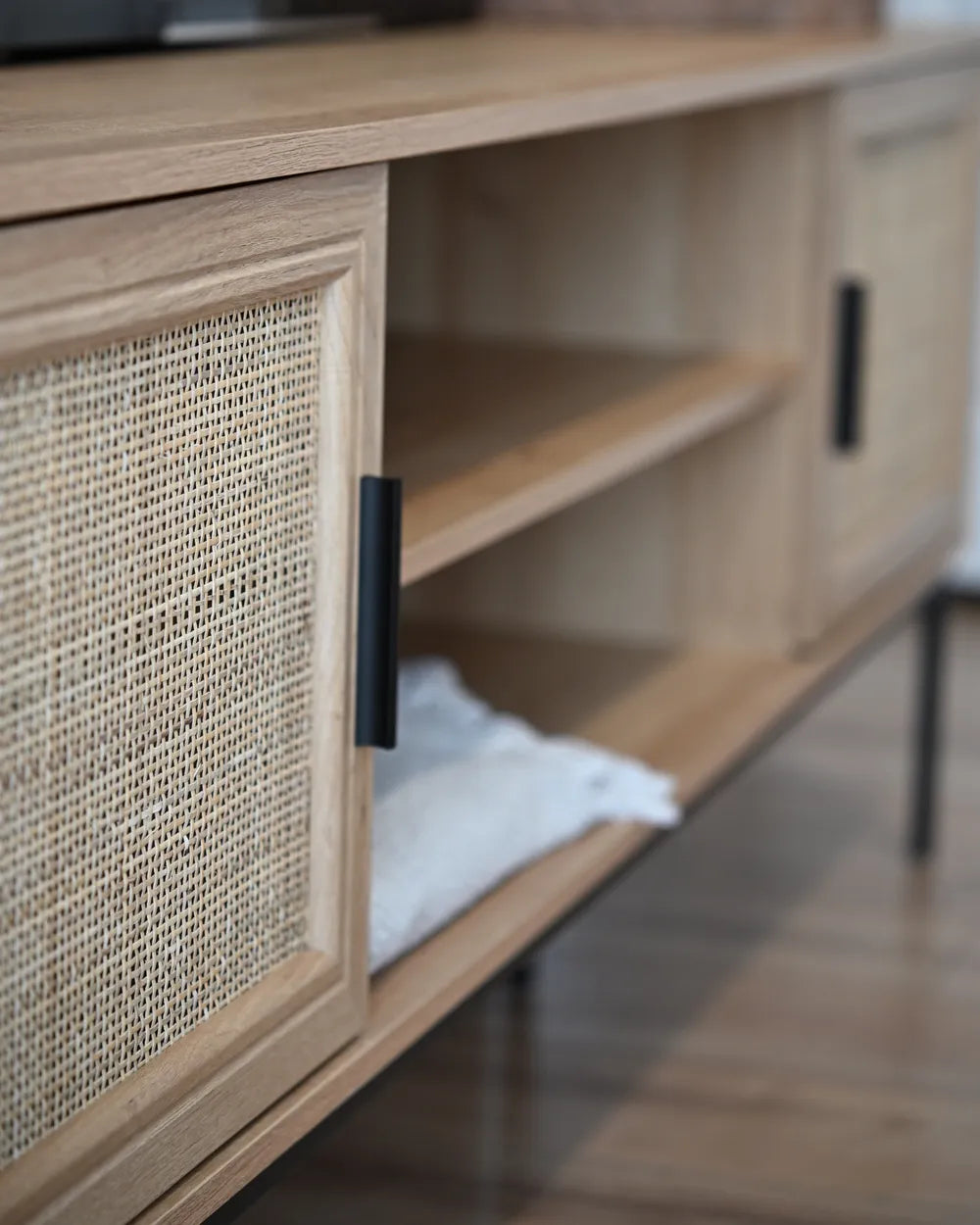 Close-up of a light wood media console with woven cane doors and black metal handles, showing open shelving and a folded white towel inside.