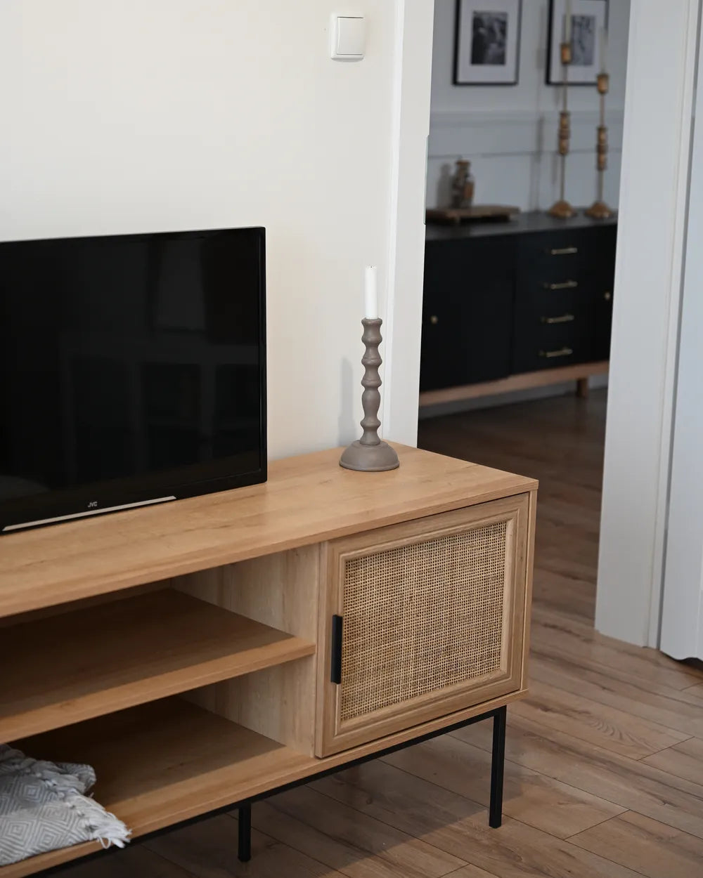 A light oak TV stand with a woven cane cabinet door, black metal legs, and open shelves, styled in a modern living room next to a black television.