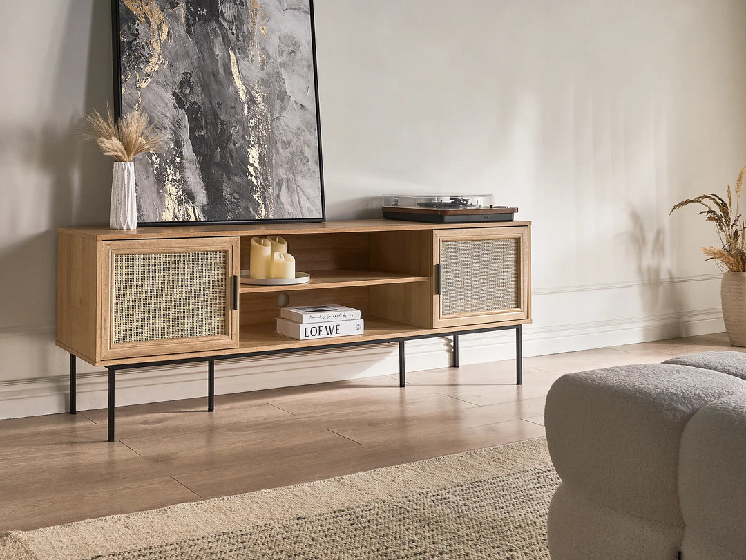 A mid-century modern wooden TV stand with woven cane doors and black metal legs, styled in a living room setting with a large framed print and decorative items.