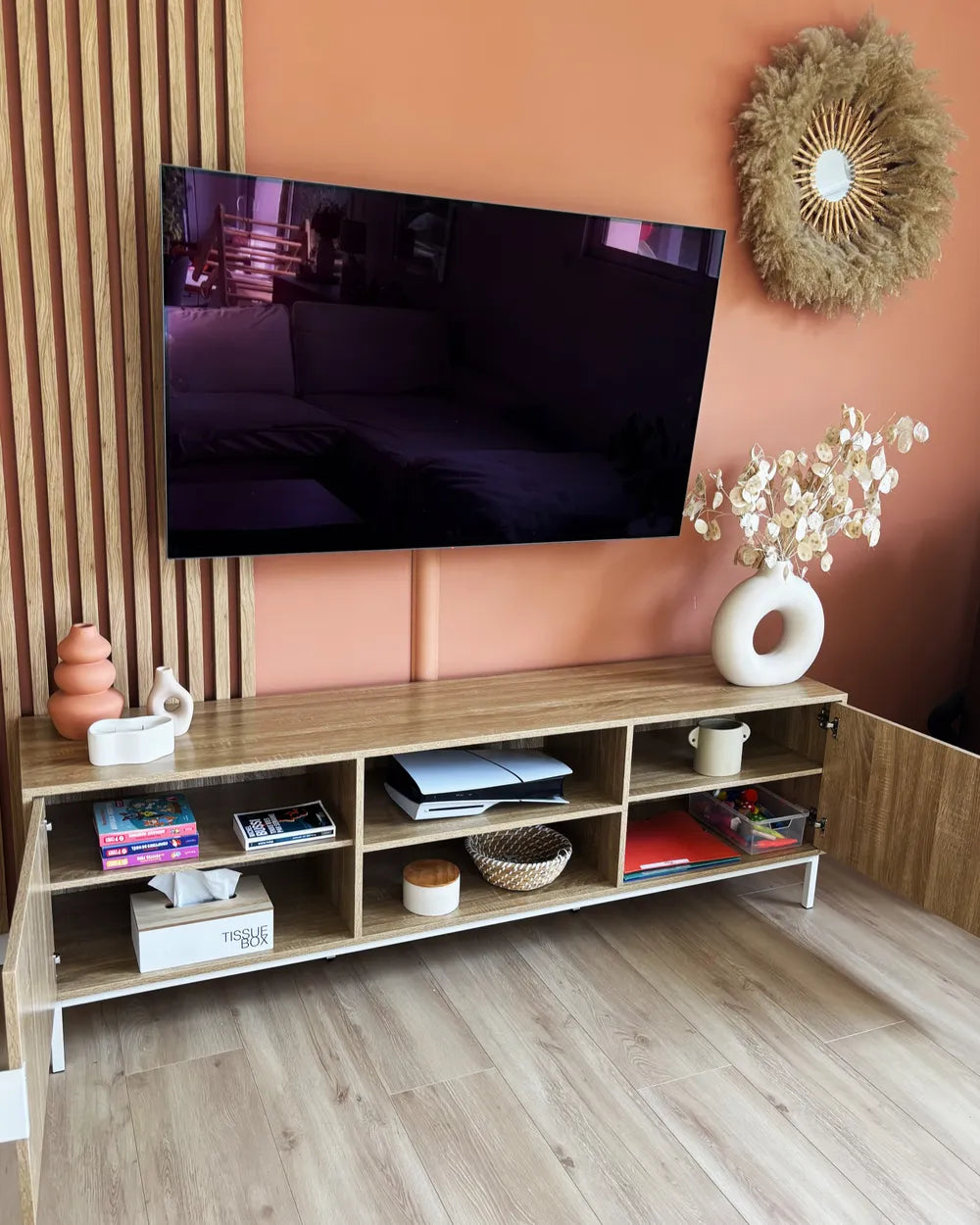 A modern light wood TV stand with open shelves and white legs, styled in a living room setting against a terracotta wall with a mounted television and decorative accessories.