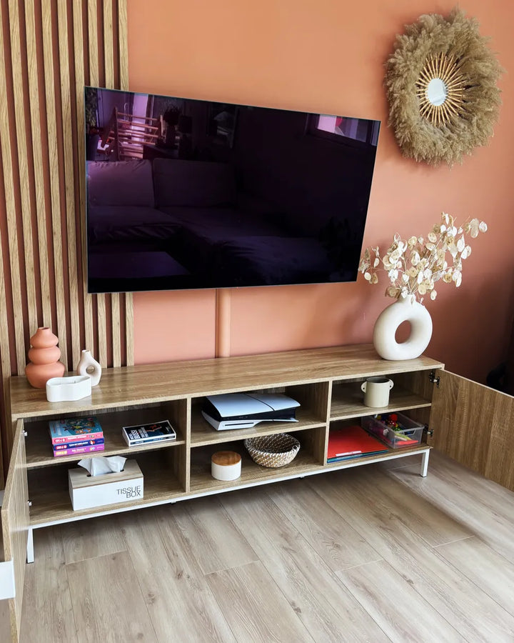 A modern light wood TV stand with open shelves and white legs, styled in a living room setting against a terracotta wall with a mounted television and decorative accessories.