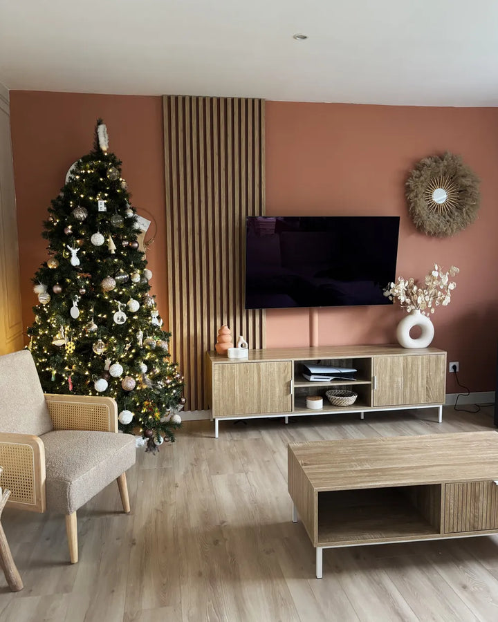 A styled living room featuring a decorated Christmas tree, a terracotta accent wall, a slatted wood panel, a wall-mounted TV, a light wood media console, a matching coffee table, and a beige armchair.