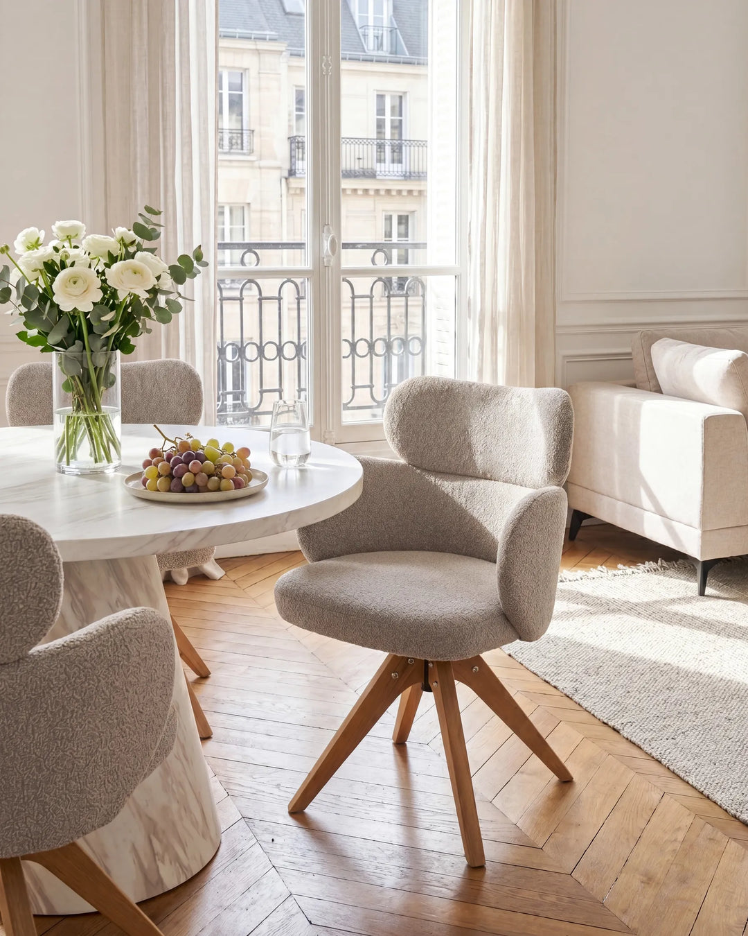 A modern dining room scene featuring a round white marble table with a glass vase of white roses and a plate of grapes, surrounded by upholstered swivel chairs in a light gray bouclé fabric with natural wood legs. The room has warm wood herringbone floors, a light beige sofa, and a large window looking out onto a Parisian balcony.
