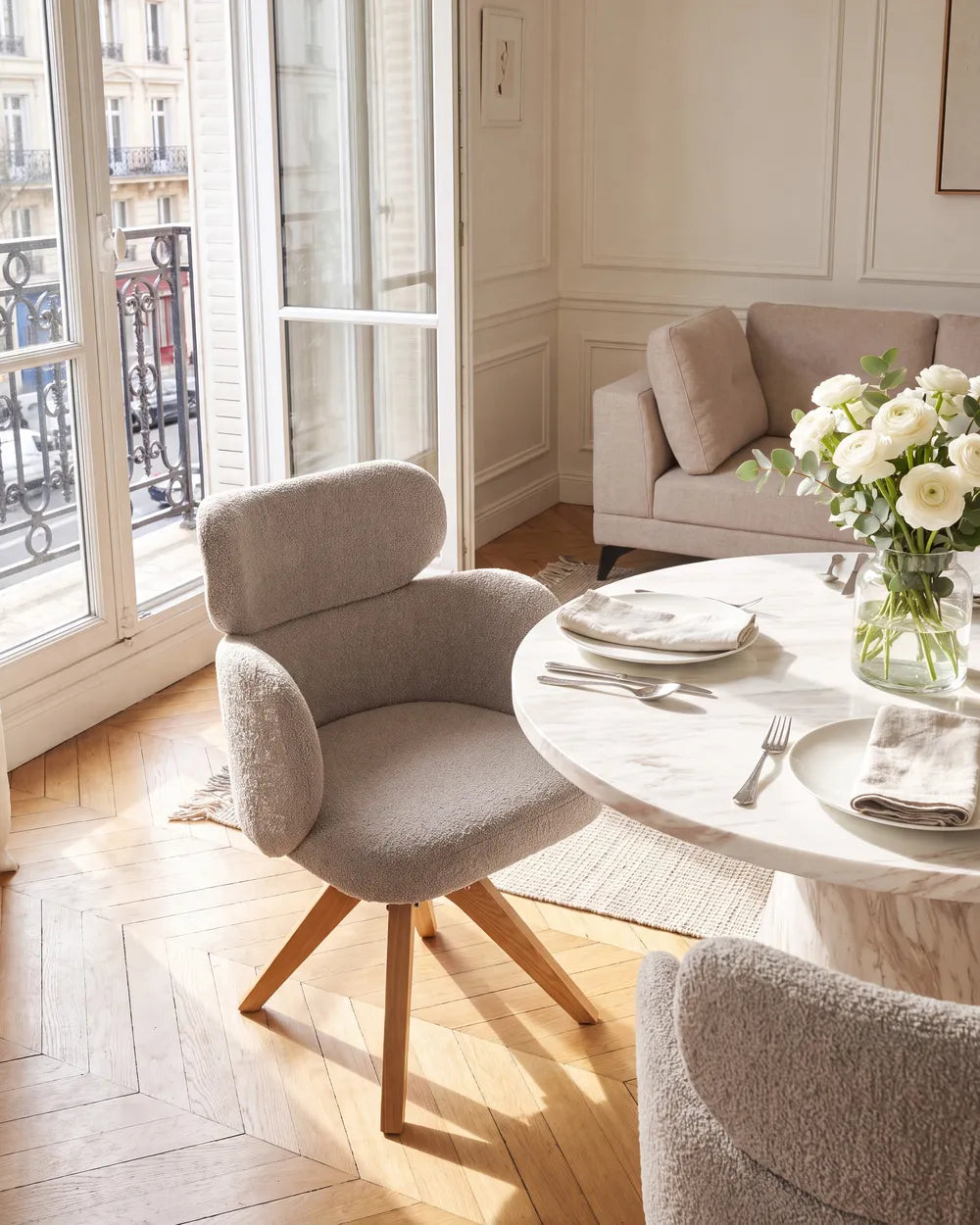 A modern upholstered dining chair in a light gray bouclé fabric with a curved back and wooden legs, shown in a bright, sunlit dining room next to a round marble table with white flowers.