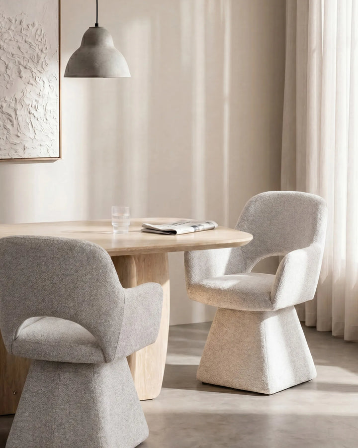 A modern dining setup featuring two sculptural light gray upholstered chairs around a round light wood table in a softly lit interior with curtains and a pendant light.