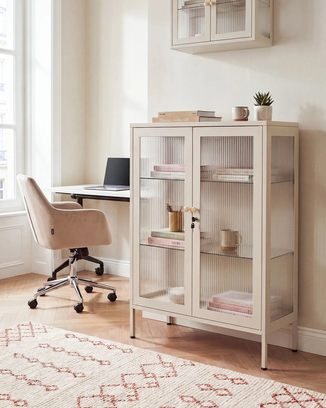 A tall cream-colored glass-front cabinet with ribbed glass doors stands in a bright home office next to a desk and beige office chair. The cabinet is styled with books, a small potted plant, and a mug on top and inside.