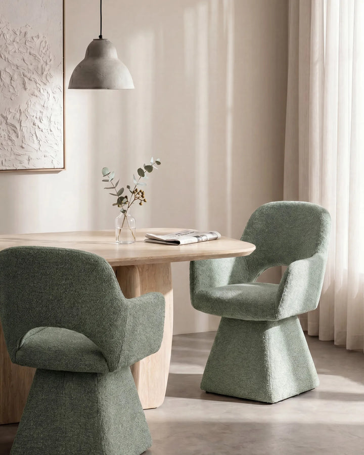 Two modern upholstered dining chairs in a muted sage green fabric arranged around a light wood table in a softly lit interior with curtains and a pendant light.