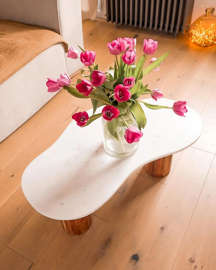 White marble coffee table with wooden legs holding a vase of pink tulips on a wooden floor.