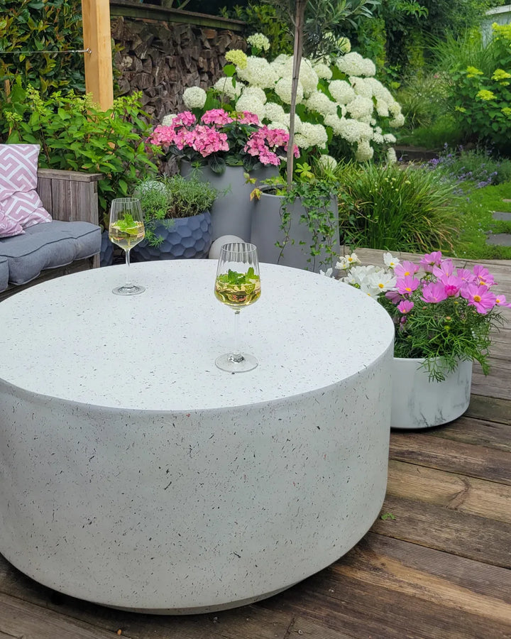 Round concrete table with two glasses of white wine on a wooden deck, surrounded by potted plants and flowers.