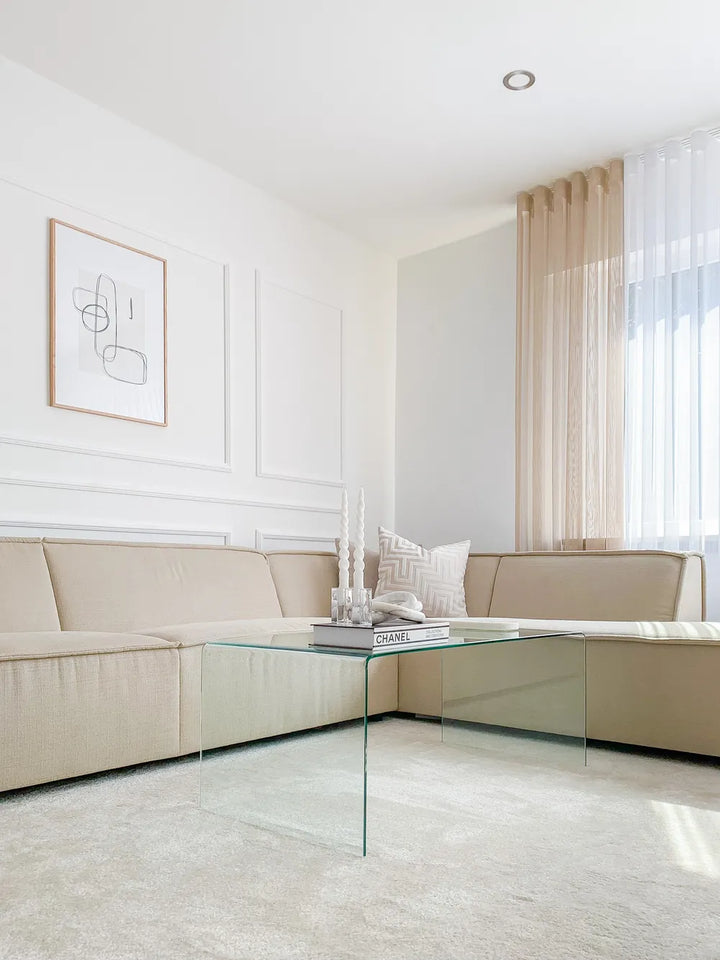 Modern living room with beige sectional sofa, glass coffee table, and abstract art on white walls.
