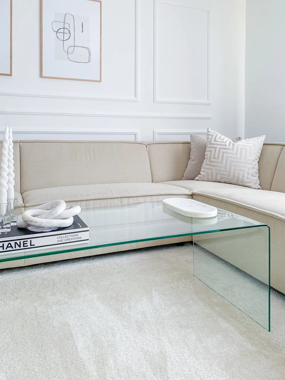 Modern living room with a glass coffee table, beige sectional sofa, and abstract art on the wall.