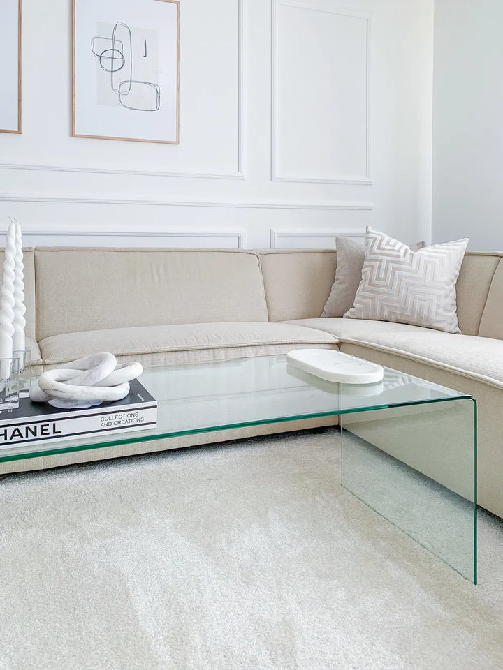 Modern living room with a glass coffee table, beige sectional sofa, and abstract art on the wall.