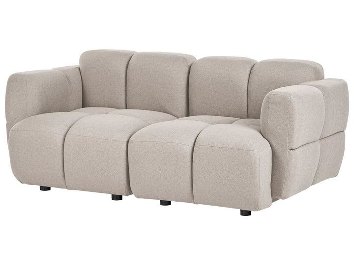 A modern modular upholstered sofa in a light beige fabric, featuring a segmented, tufted design with blocky arms and dark cylindrical legs, shown against a plain white background.